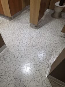 Public restroom with wooden stall doors and a mosaic white-and-gray tile floor; a toilet is visible in the back. LVT Tile clean and reseal