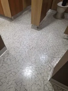 Public restroom with wooden stall doors and a mosaic white-and-gray tile floor; a toilet is visible in the back. LVT Tile clean and reseal