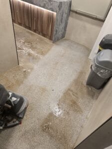 A small toilet area with wooden cabinet doors and a concrete-style countertop, and a wet, stained safety floors.