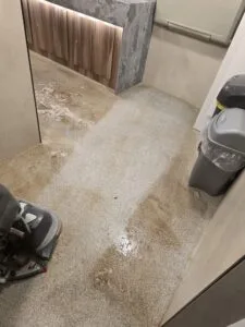 A small toilet area with wooden cabinet doors and a concrete-style countertop, and a wet, stained safety floors.